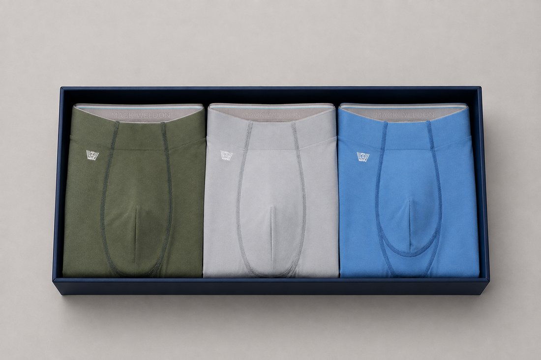 Three pairs of folded men's underwear in olive green, light gray, and blue, neatly arranged inside a dark navy blue presentation box on a light gray background.