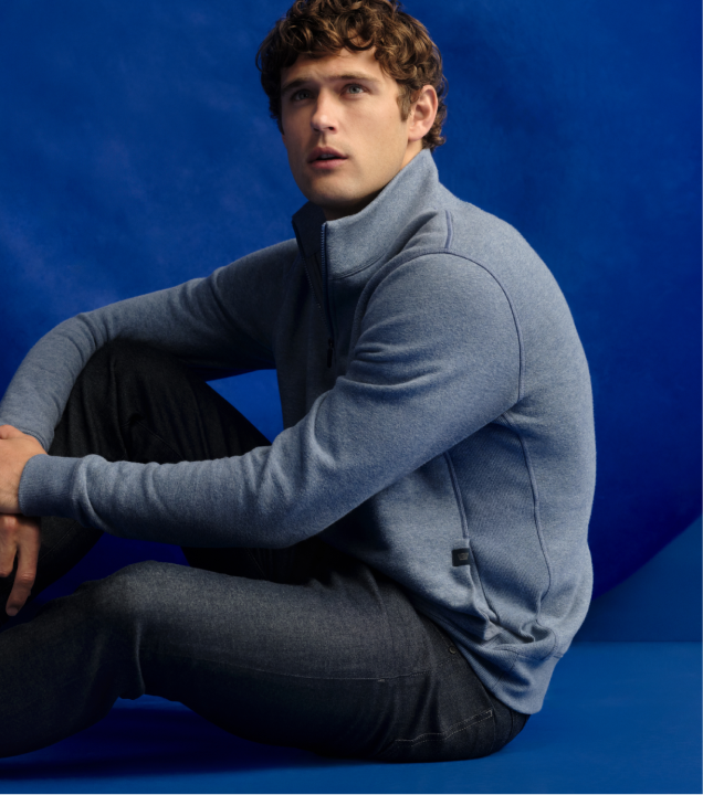 Man sits on ground with blue backdrop. He is wearing jeans and an ACE half zip