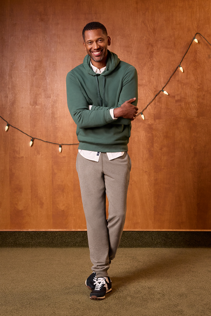Man wearing a green sweater and gray pants standing against a wooden wall with string lights.
