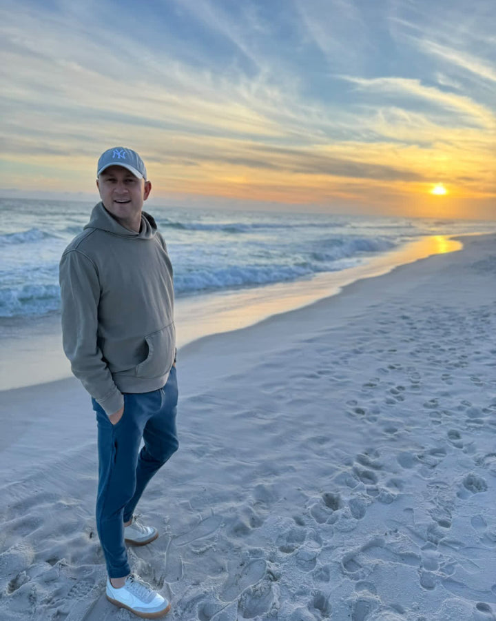 Man stands on the beach with a sunset behind him. He is wearing a Mack Weldon Hoodie and Mack Weldon sweatpants