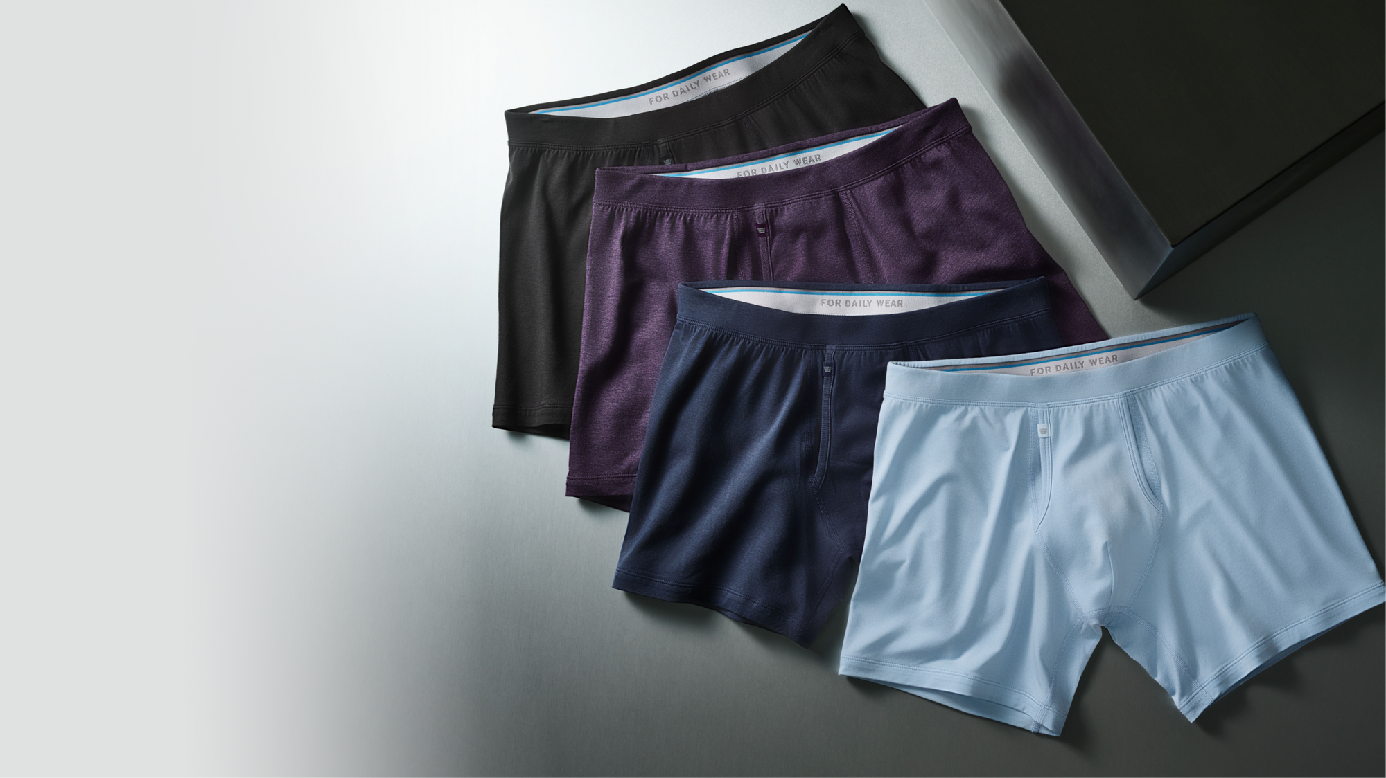 Four pairs of men's boxer briefs in black, purple, navy blue, and light blue on a grey surface.