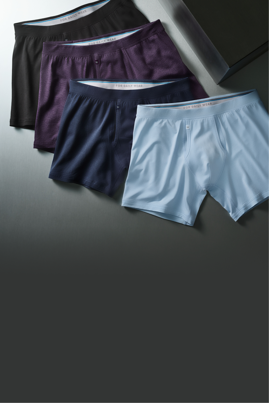 Four pairs of men's boxer briefs in black, purple, navy blue, and light blue on a grey surface.