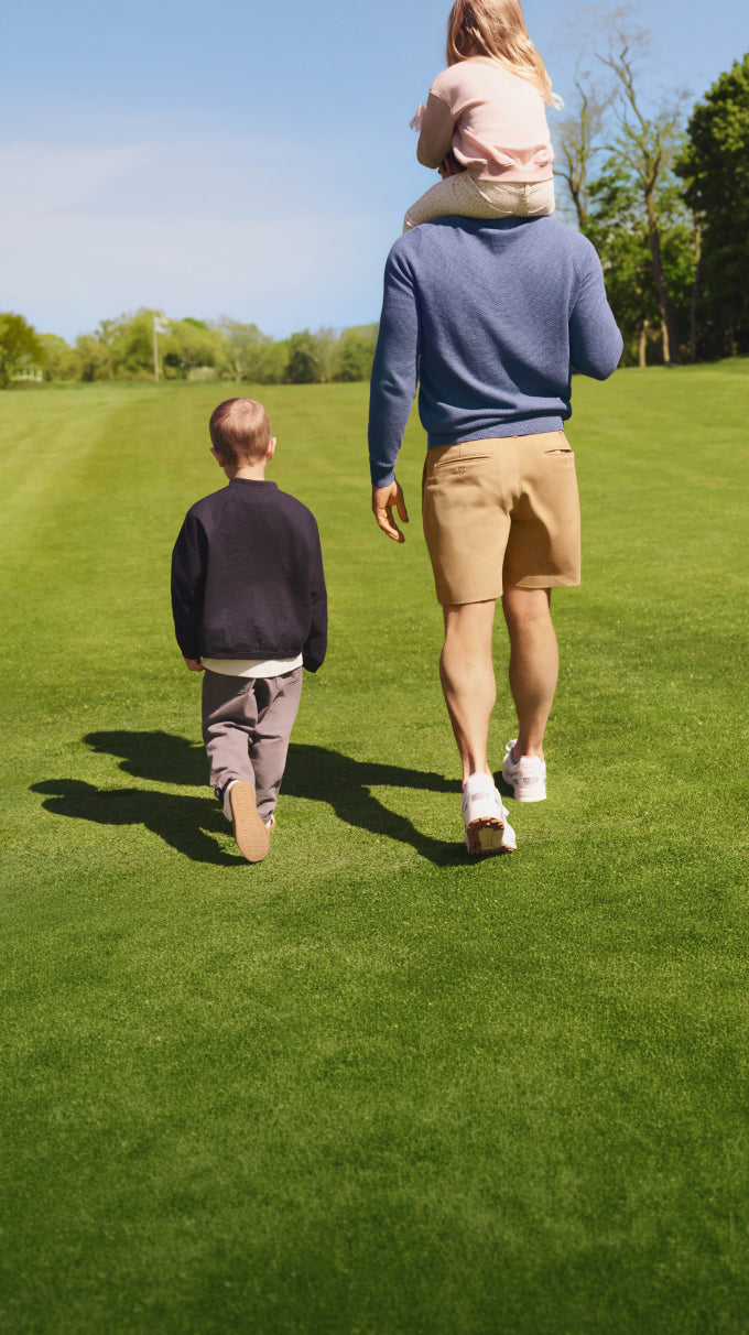 A man walking on a grassy lawn with his two children, carrying a young girl on his shoulders while a boy walks alongside them.