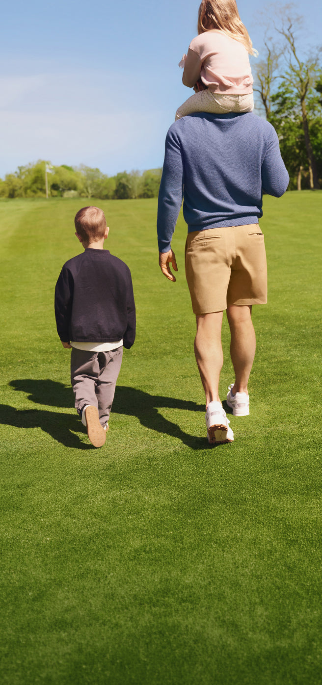 A man walking on a grassy lawn with his two children, carrying a young girl on his shoulders while a boy walks alongside them.