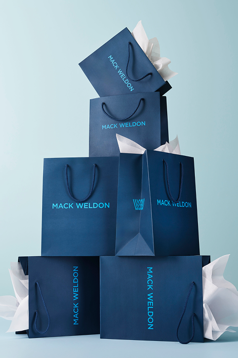Stack of blue 'Mack Weldon' shopping bags on a light blue background