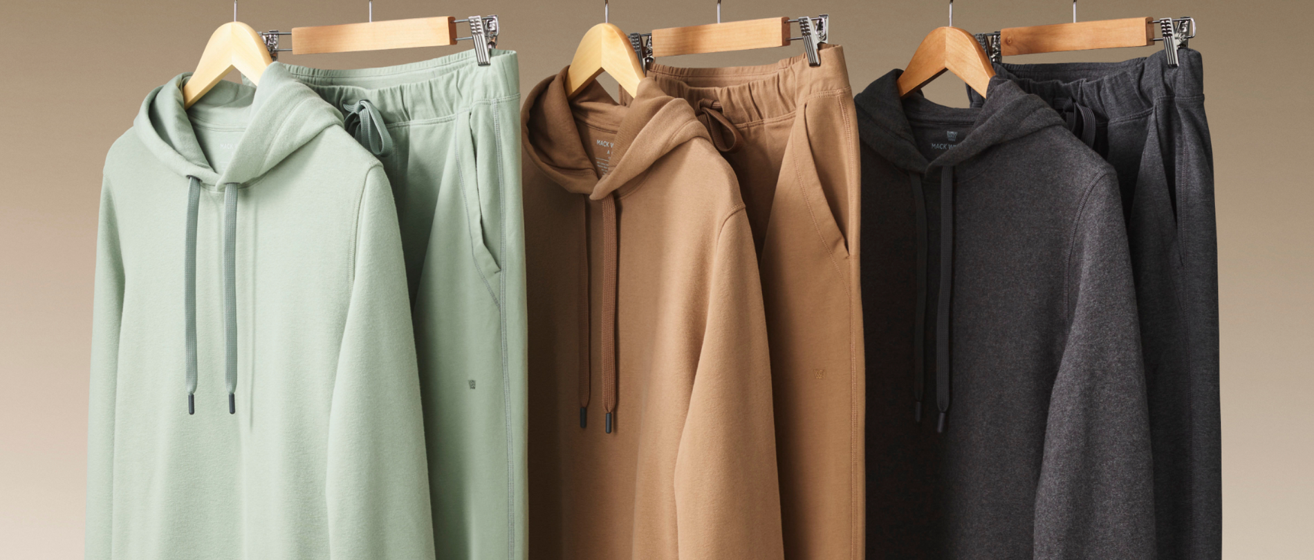 Four hoodies in different colors hanging on wooden hangers against a beige background