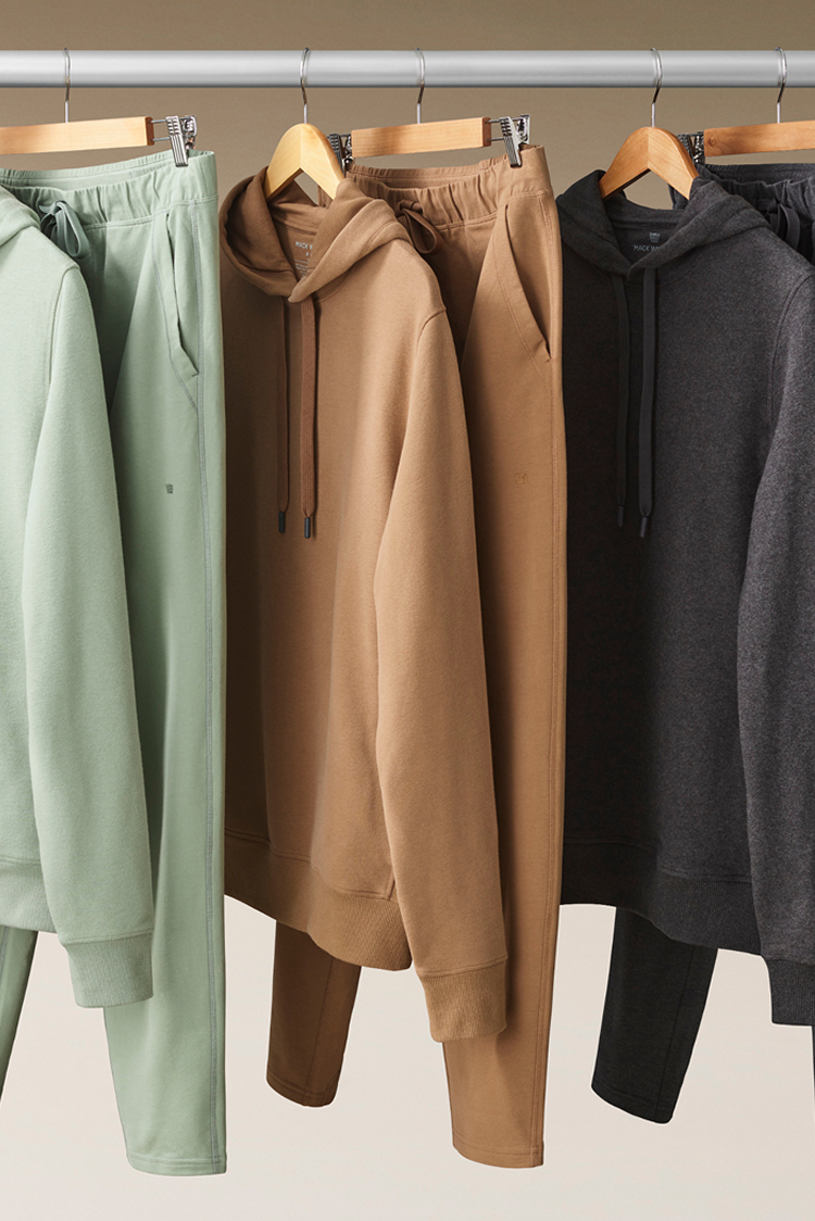 Three hoodies in green, brown, and black hanging on a rack against a neutral background.