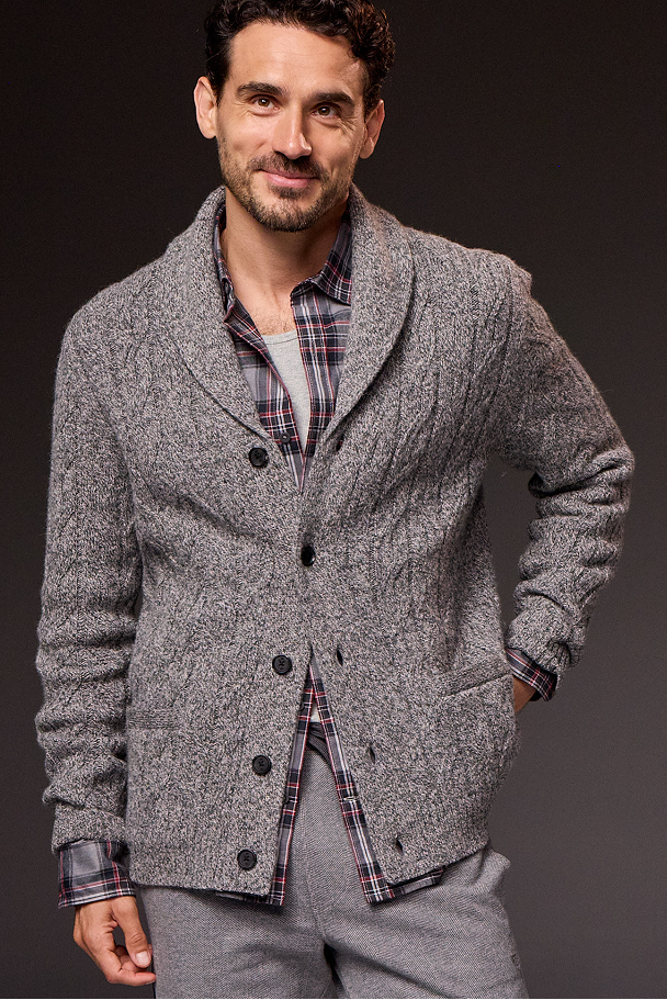 Man wearing a gray textured cardigan over a plaid shirt with a dark background