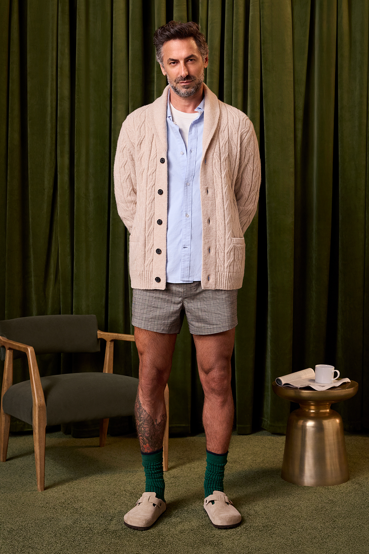 Man wearing a beige cardigan, light blue shirt, gray shorts, and green socks standing in front of green curtains.