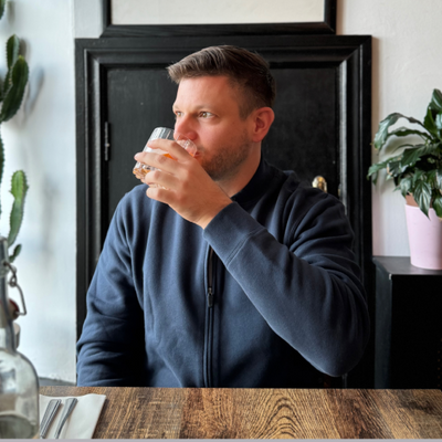 Man wearing Mack Weldon Ace Bomber Jacket sitting in wooden table, taking a sip from a glass.