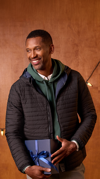 Man wearing a black puffer jacket with a green hood, holding a wrapped gift against a wooden background.