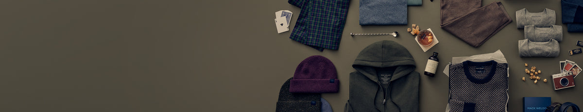 A styled flat-lay of various men's clothing items, including a dark gray hoodie, a purple knit beanie, a blue plaid shirt, a gray t-shirt, and folded garments, arranged on a dark gray background.